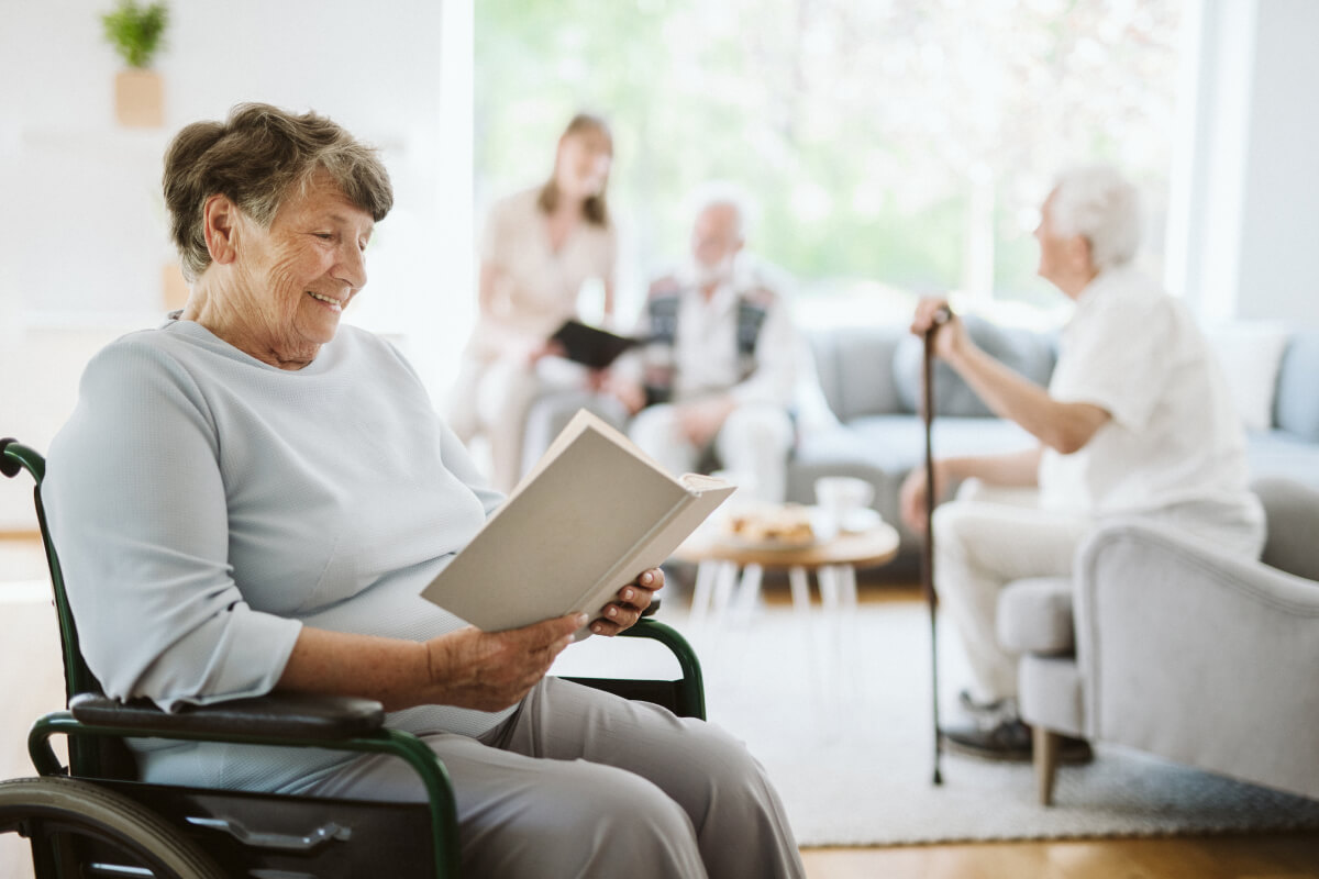 A nurse in white scrubs warmly embraces an elderly woman on a sofa, both smiling. The setting is bright and cozy, conveying a sense of care and happiness.