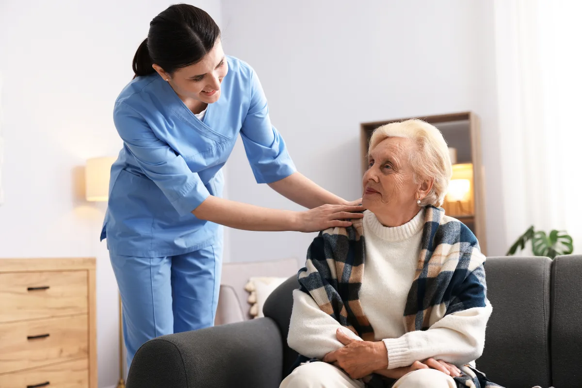 A nurse in blue scrubs gently places a hand on an elderly woman's shoulder, who is seated on a sofa wrapped in a plaid blanket, conveying warmth and care.