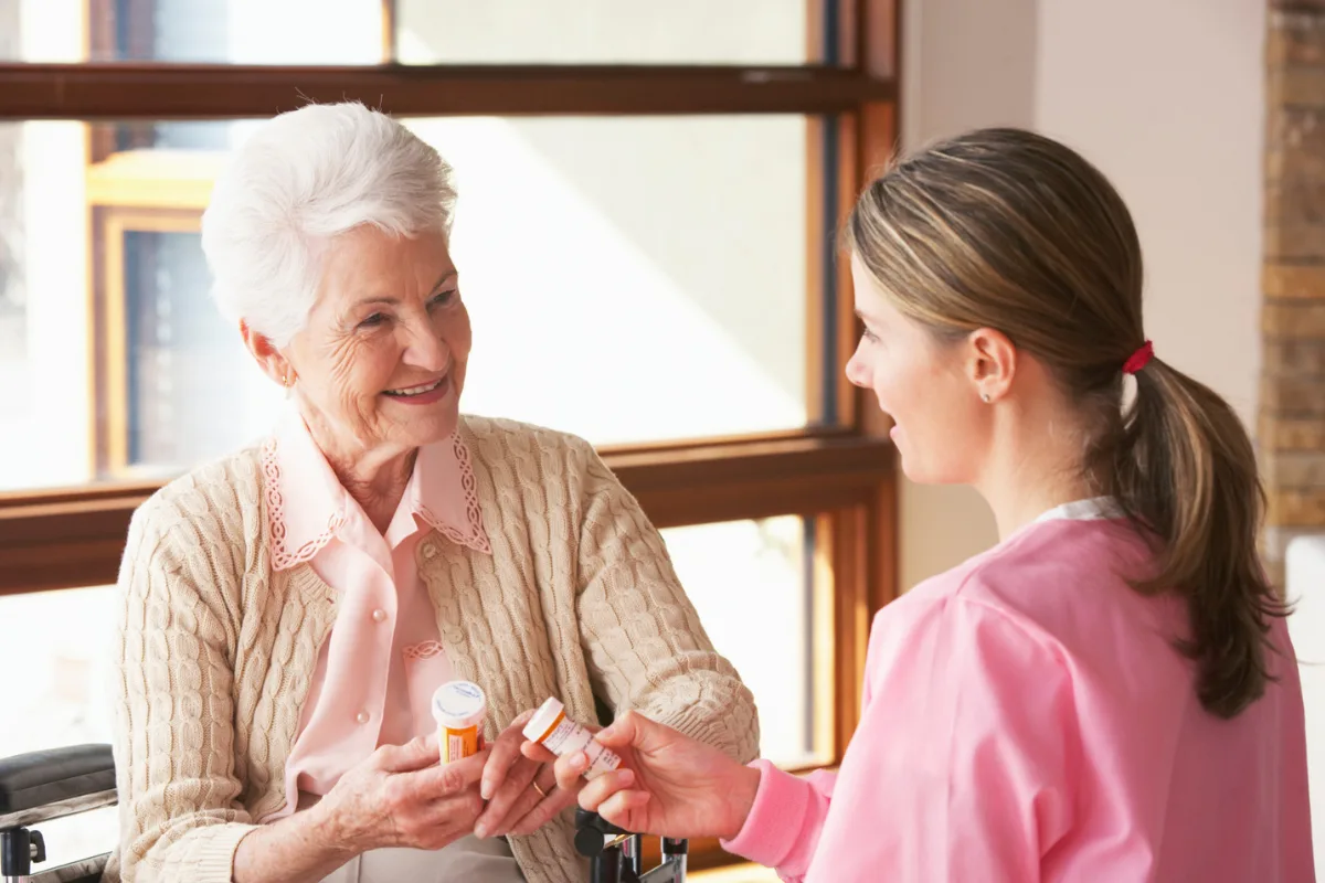 A nurse in pink scrubs assists a smiling elderly woman in a beige cardigan with medication, conveying care and support in a bright, sunlit room.