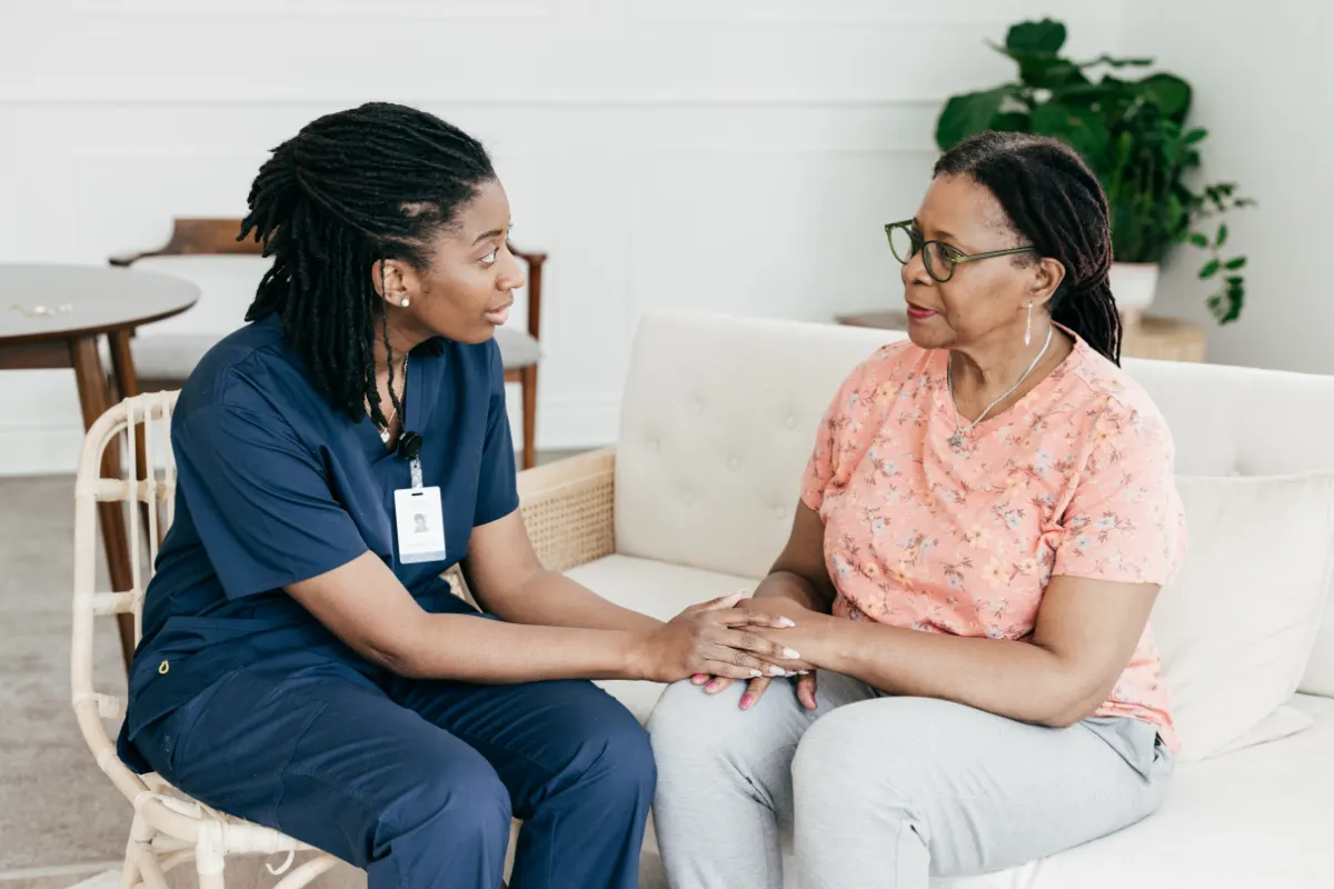 A nurse in blue scrubs holds an elderly woman's hands on a sofa, conveying empathy and care. The setting is calm, with plants in the background.