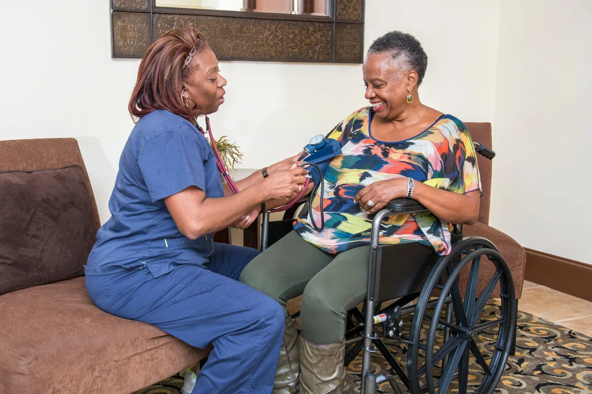 A nurse in blue scrubs checks the blood pressure of a smiling woman in a wheelchair, creating a warm, caring atmosphere in a cozy room.