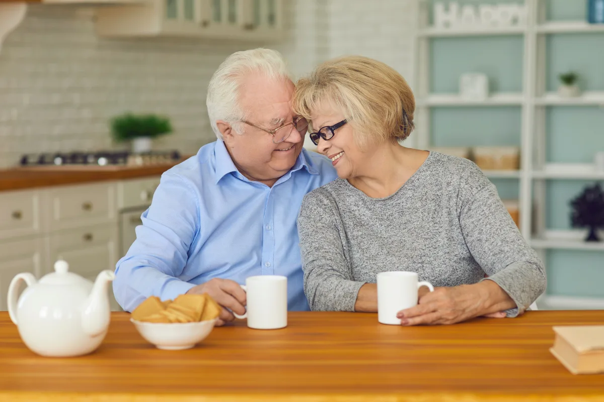 Elderly couple sitting at a kitchen table, smiling warmly at each other, holding mugs. A teapot and cookies are on the table, creating a cozy atmosphere.