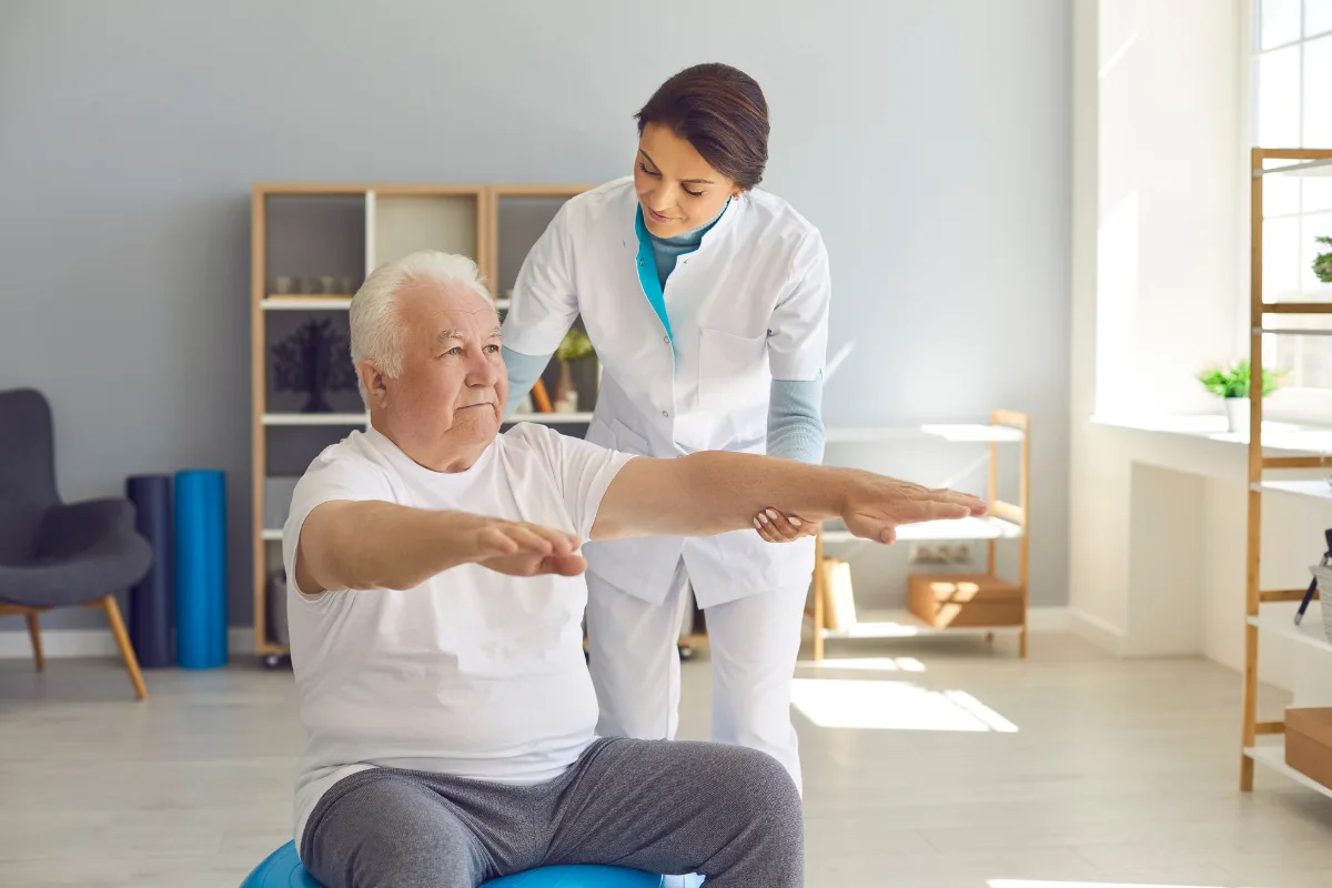 A senior man sits on a blue exercise ball, arms extended. A healthcare professional assists him in a bright, modern room, conveying support and focus.