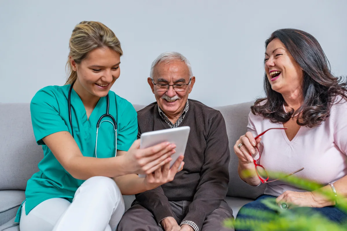 A nurse in teal scrubs shows a tablet to a smiling elderly man and woman on a couch. They seem engaged and happy, conveying a positive atmosphere.