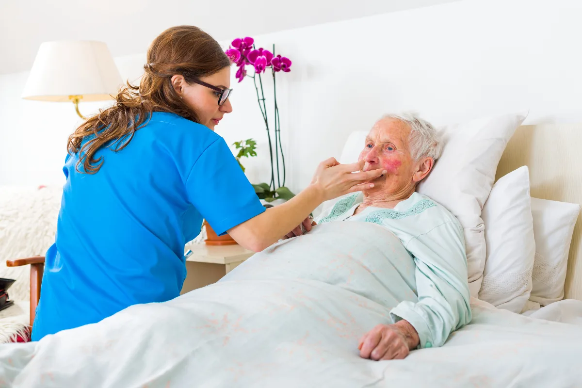 A caregiver in blue scrubs gently feeds an elderly person resting in bed, with white bedding and pink flowers nearby. The mood is caring and nurturing.