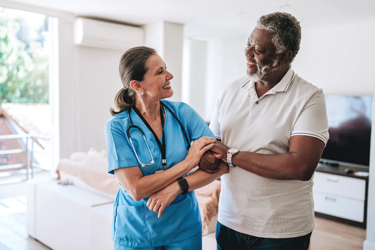 A smiling female nurse in blue scrubs and a stethoscope warmly interacts with a cheerful male patient in a living room, conveying care and positivity.