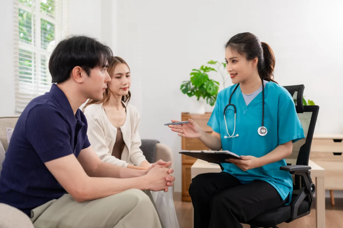 A doctor in blue scrubs holds a clipboard, attentively consulting a seated couple in an office. The room is bright, with plants in the background.