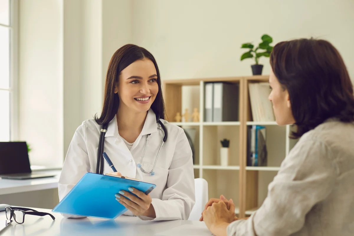 A smiling female doctor with a stethoscope holds a clipboard while consulting with a patient in a bright office, creating a warm, professional atmosphere.