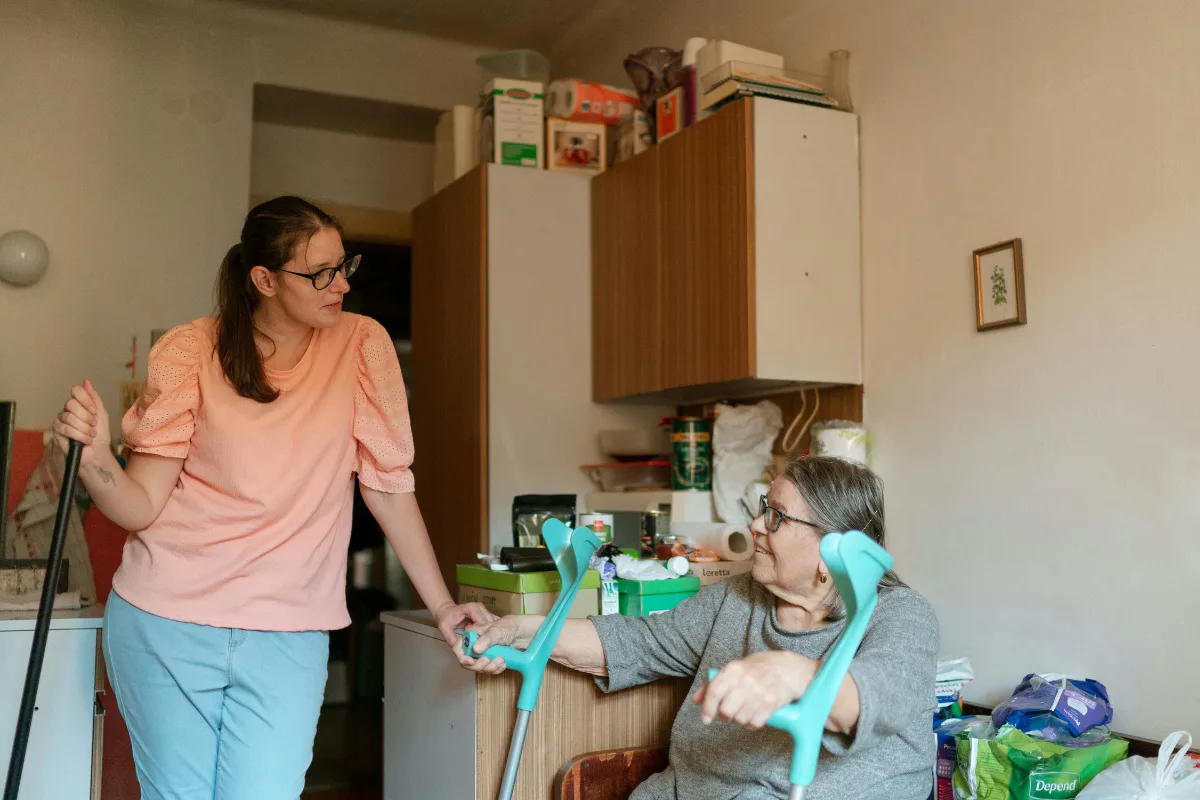 A young woman in a peach shirt leans on a cane, speaking to an elderly woman with crutches in a cluttered room, suggesting care and support.