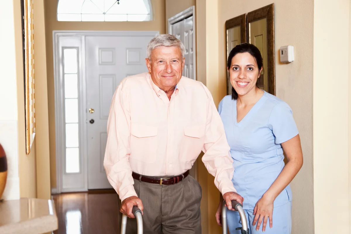 Elderly man using a walker stands by a smiling caregiver in a blue uniform in a bright hallway, conveying support and companionship.