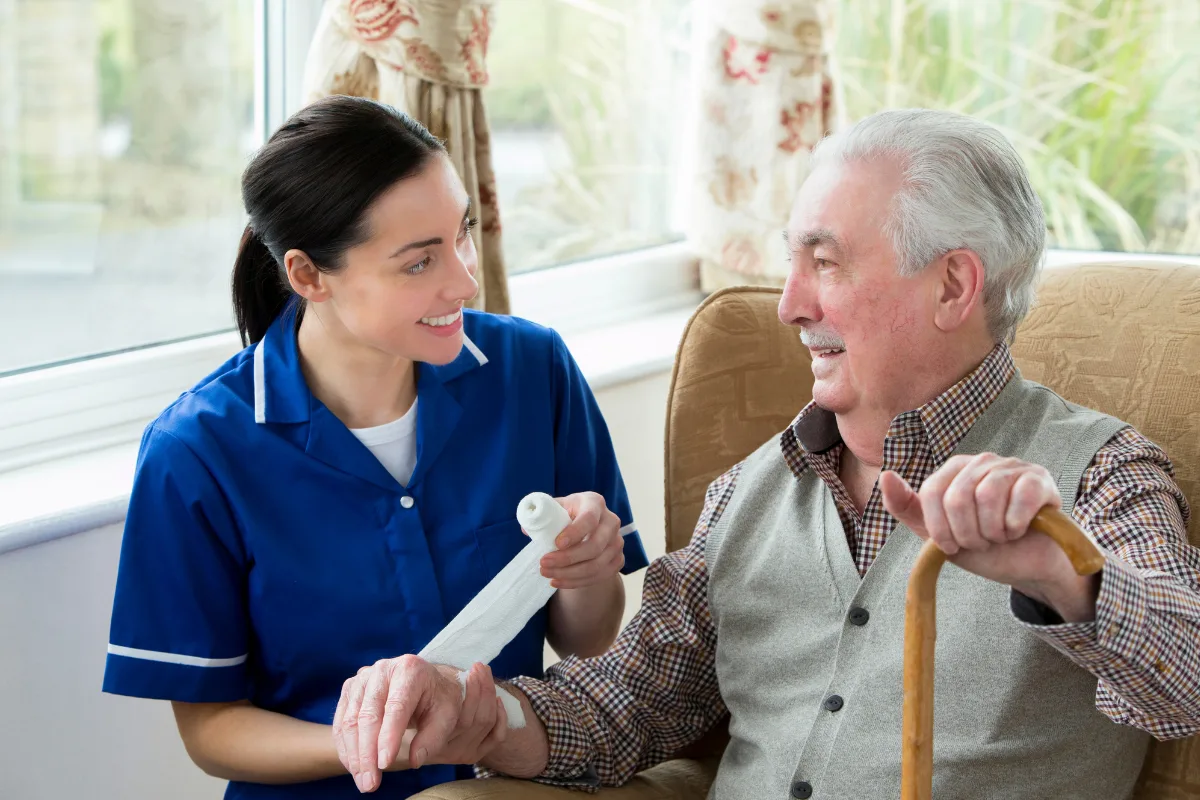 A nurse in a blue uniform bandages an elderly man's hand as they both smile warmly. The man sits in a chair with a cane, conveying a caring, supportive mood.
