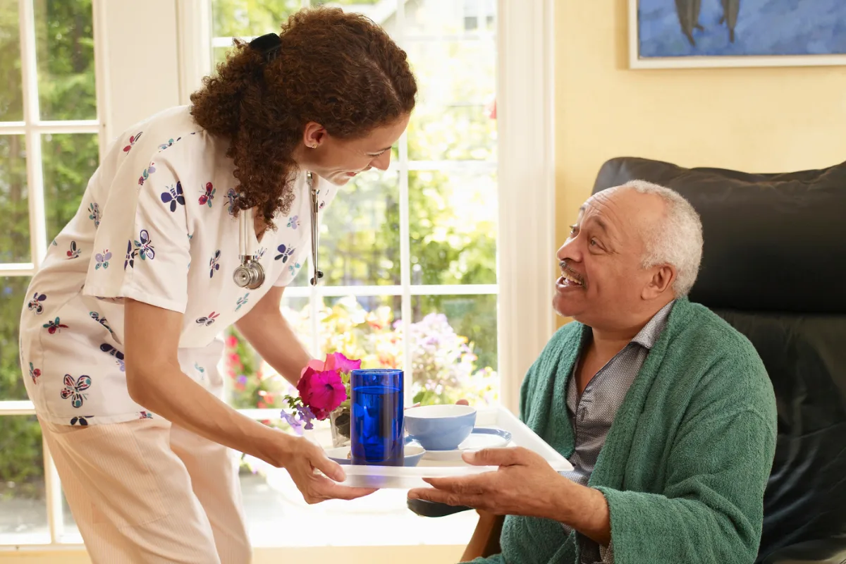 A caregiver in a floral uniform hands a meal tray to a smiling elderly man in a green robe. The room is bright with a cheerful, caring atmosphere.