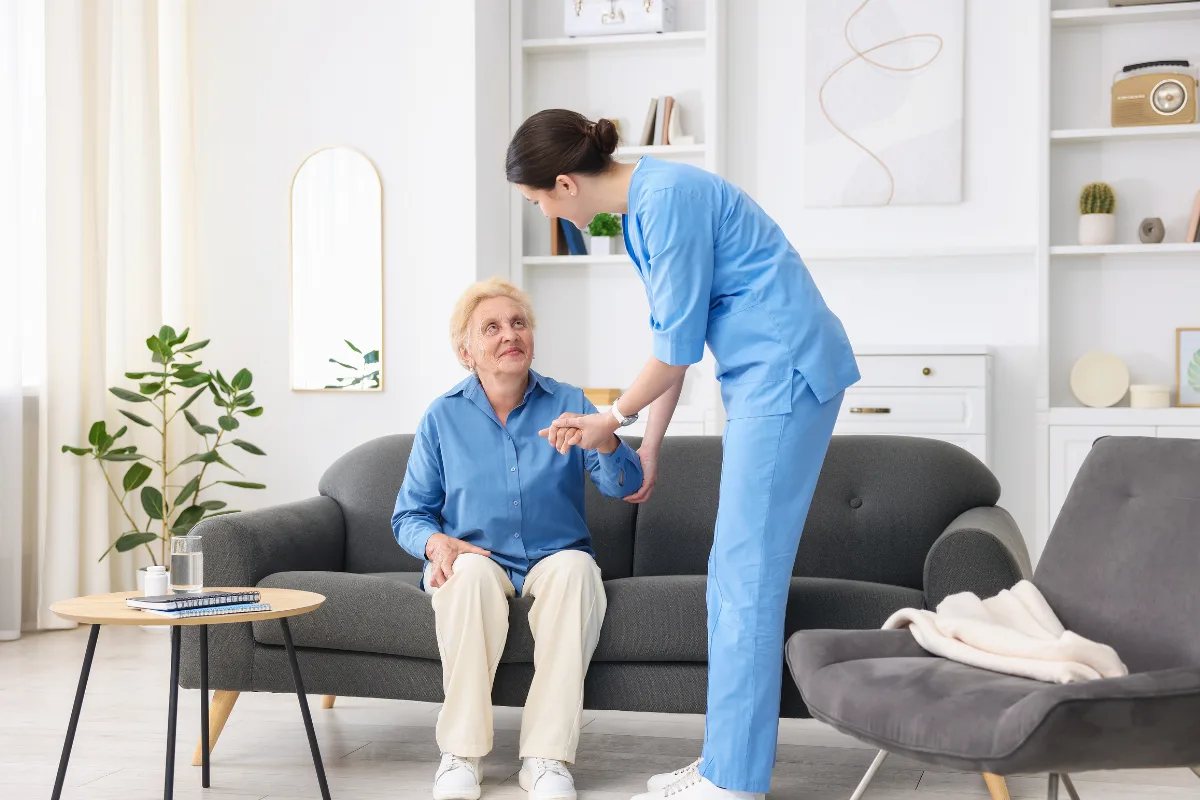 A caregiver in blue scrubs helps an elderly woman stand from a sofa in a bright living room. The atmosphere is supportive and caring.