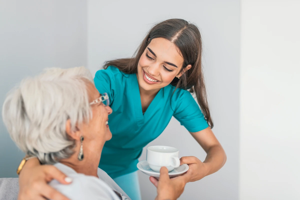 A smiling young caregiver in a turquoise uniform offers a cup of tea to an elderly woman with white hair, creating a warm and compassionate interaction.