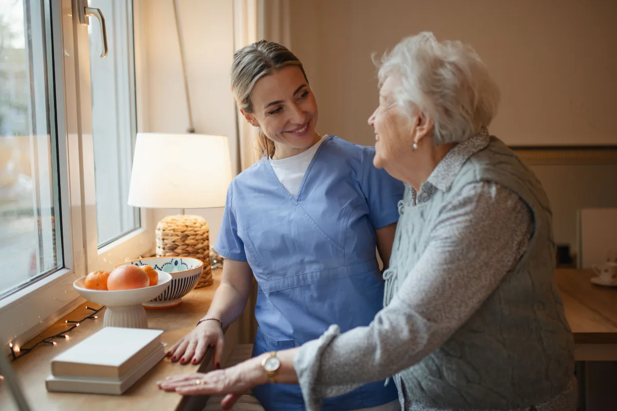 A nurse in blue scrubs smiles warmly at an elderly woman in a gray vest, standing by a window with a lamp and a bowl of oranges, conveying care and companionship.