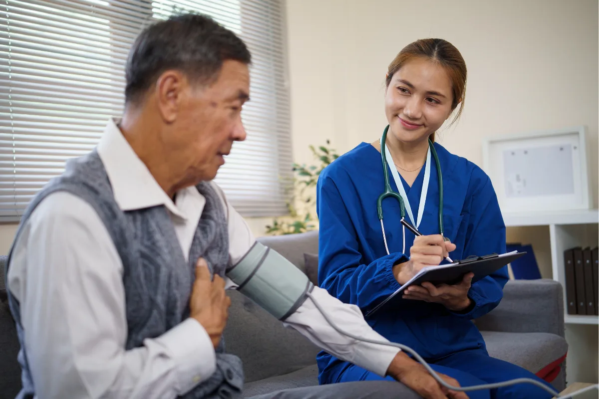 A nurse in blue scrubs smiles while checking an elderly man's blood pressure. She holds a clipboard, conveying a caring, professional atmosphere.