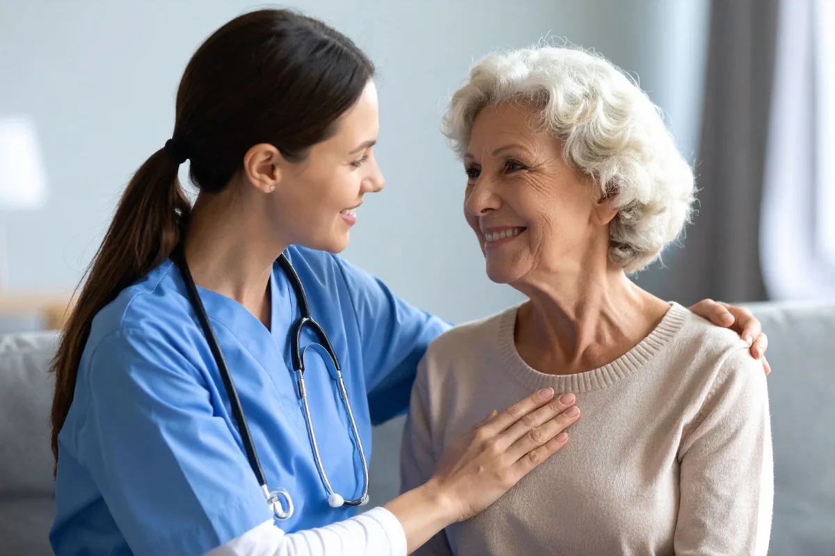A smiling nurse in blue scrubs and stethoscope gently places a hand on an elderly woman's shoulder, who smiles back warmly in a caring environment.
