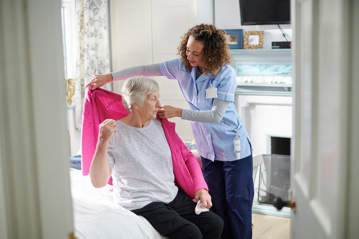 A caregiver in a blue uniform smiles warmly at an elderly woman in a pink sweater while bandaging her hand. They sit in a cozy, bright room.