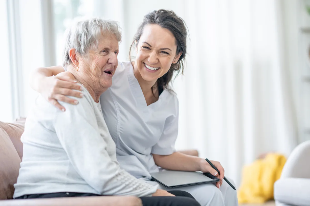 A nurse and an elderly woman share a joyful moment on a couch, smiling warmly. The setting is bright and homey, conveying care and companionship.