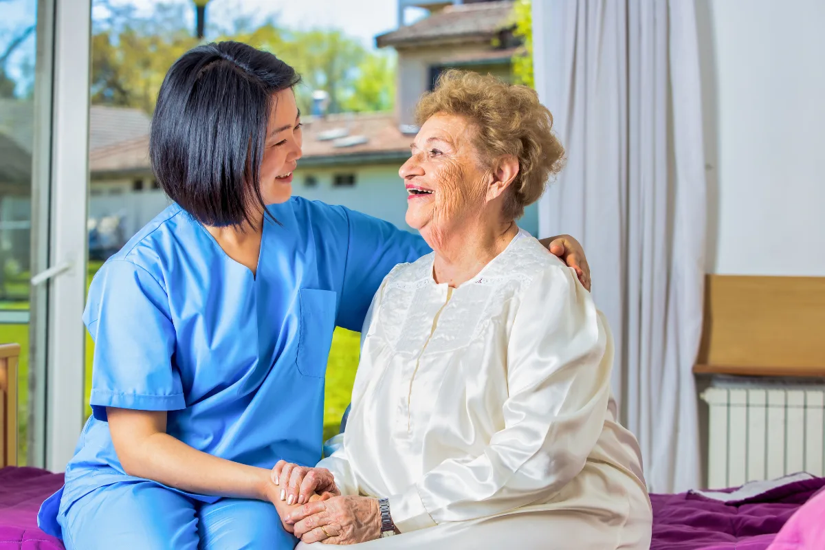 A nurse in blue scrubs and an elderly woman in a white gown share a warm, joyful moment. They sit on a bed by a window, smiling and holding hands.