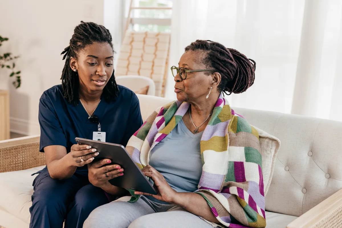 A nurse in blue scrubs sits with an elderly woman wrapped in a colorful blanket, discussing something on a tablet. They are seated on a beige sofa.