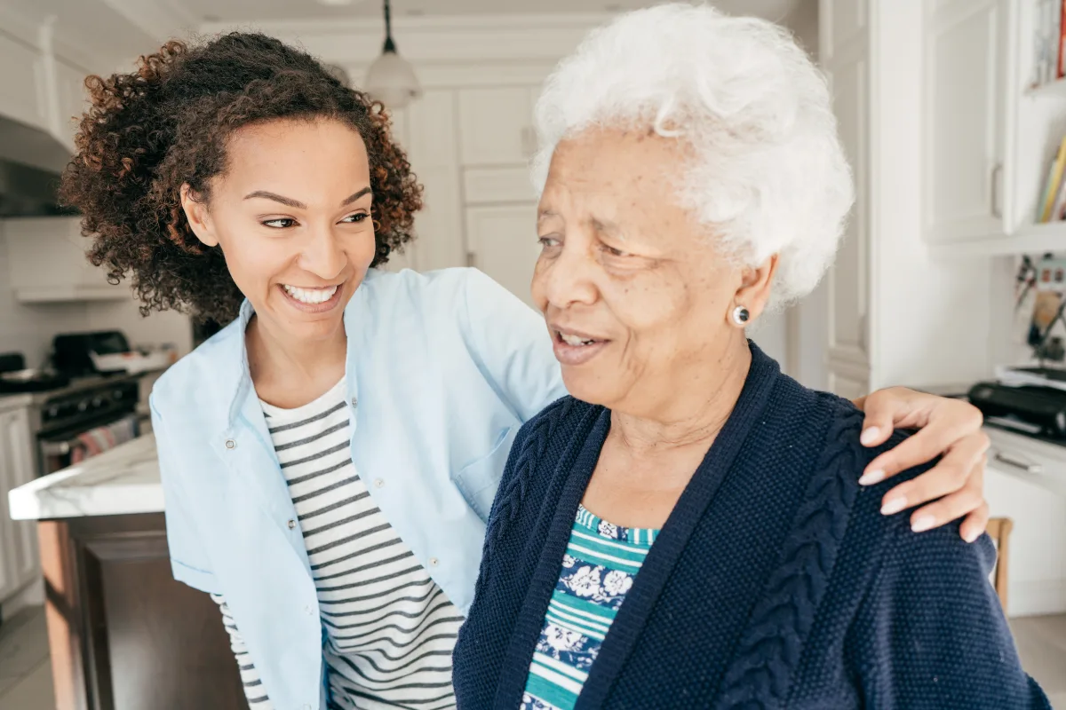 A young woman embraces an elderly woman in a bright kitchen, both smiling warmly. The scene conveys warmth, care, and family bonding.