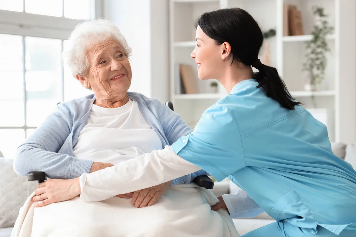 A smiling elderly woman sits in a wheelchair, kindly interacting with a young caregiver in blue scrubs. The room is bright and airy, creating a warm, caring atmosphere.