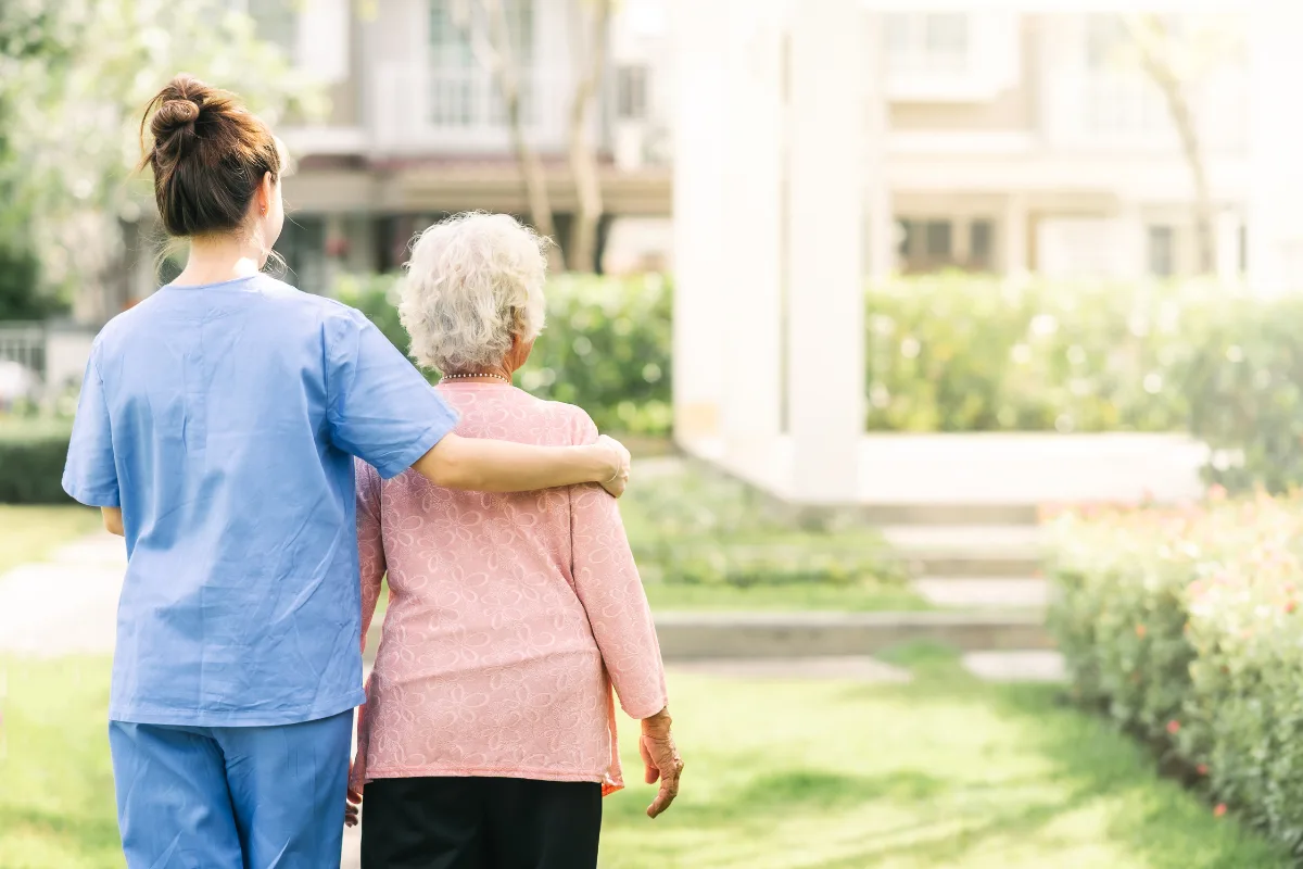 A caregiver in blue scrubs gently supports an elderly woman in a pink sweater as they walk through a sunny garden, symbolizing care and companionship.