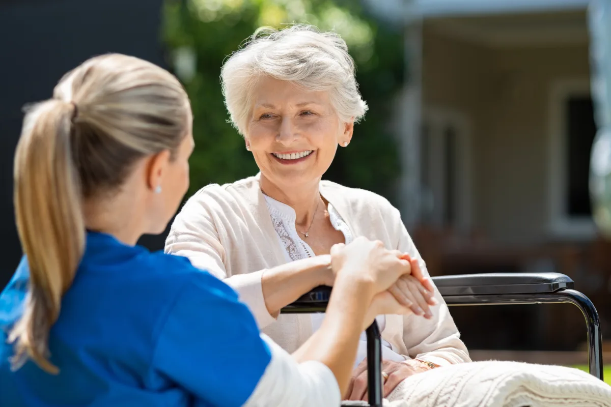Elderly woman in a wheelchair smiling at a caregiver in blue scrubs, holding hands outdoors. Warm, caring interaction in a peaceful setting.