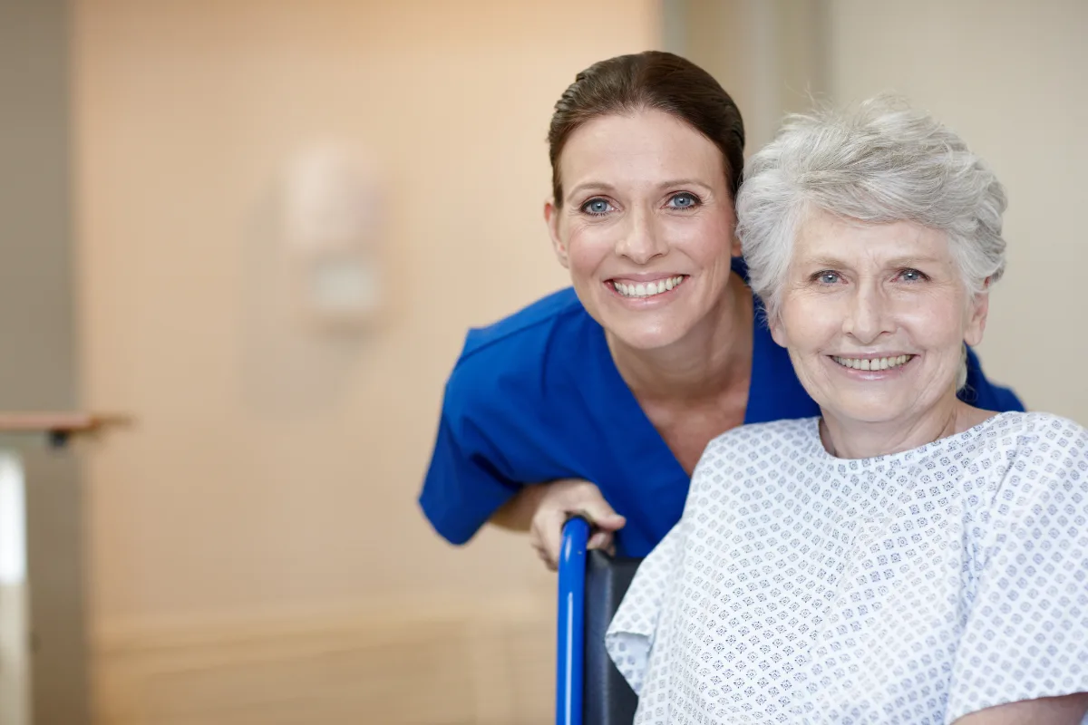 A cheerful nurse in blue scrubs leans next to a smiling elderly woman in a hospital gown, conveying warmth and care in a medical setting.