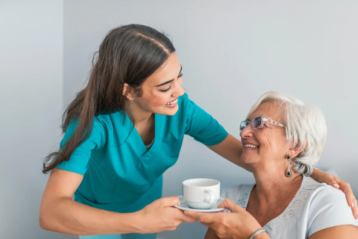 A young caregiver in a turquoise uniform hands a cup of tea to a smiling elderly woman in white. The scene conveys warmth and care.