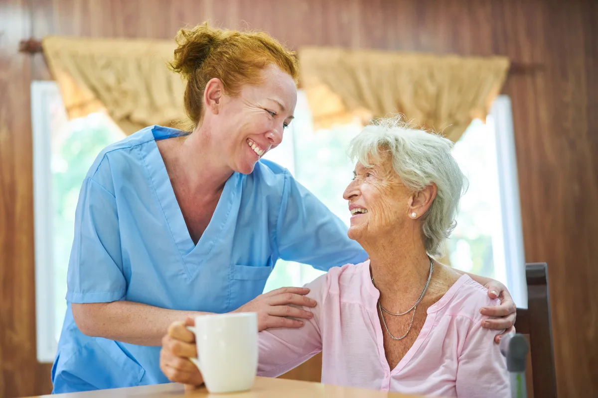 A caregiver in blue scrubs and an elderly woman share a warm smile while the woman holds a mug. The scene conveys warmth and companionship.
